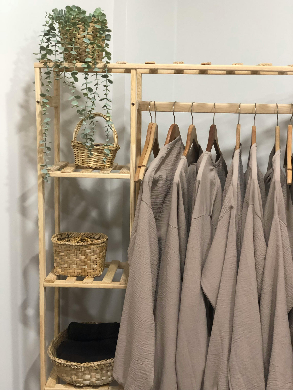 Wooden clothing rack with shelves, holding woven baskets, greenery, and neutral-toned garments for home organization.
