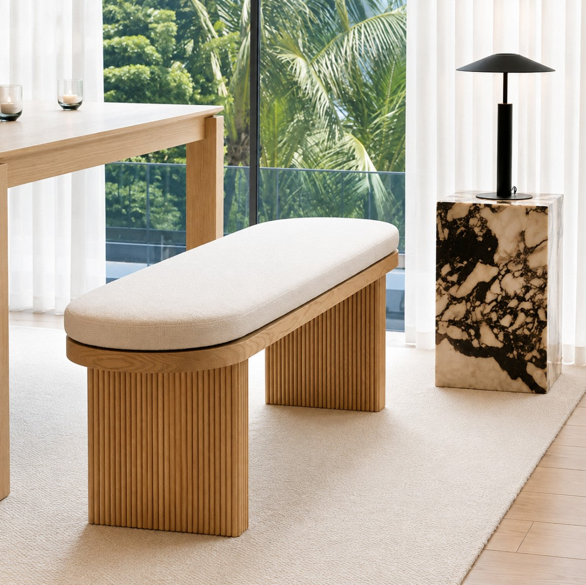 A modern wooden bench with a curved upholstered seat cushion in a bright, contemporary room. The bench features a fluted wood frame and is positioned on a light beige rug next to a marble side table with a black lamp, with a large window showing lush green tropical foliage in the background.