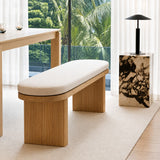 A modern wooden bench with a curved upholstered seat cushion in a bright, contemporary room. The bench features a fluted wood frame and is positioned on a light beige rug next to a marble side table with a black lamp, with a large window showing lush green tropical foliage in the background.