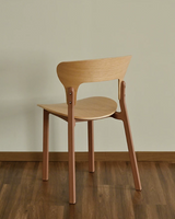 A modern wooden dining chair with a light natural wood seat and backrest, supported by four reddish-brown legs, shown from a three-quarters angle on a dark wood floor against a beige wall.