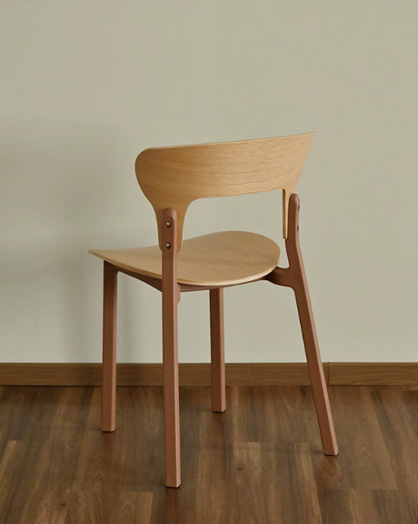 A modern wooden dining chair with a light natural wood seat and backrest, supported by four reddish-brown legs, shown from a three-quarters angle on a dark wood floor against a beige wall.
