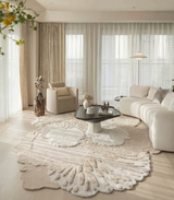 A styled living room featuring a large, organic-shaped cream shag rug with a textured, raised floral or leaf-like pattern. The room is decorated with a cream sectional sofa, a matching armchair, a black coffee table, and sheer white curtains.