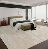 A large rectangular area rug with a distressed, textured cream and off-white pattern, styled in a modern bedroom setting with a bed, armchair, and large windows.