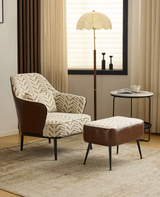 A mid-century modern armchair with a brown leather side panel and a matching ottoman, both upholstered in a beige and brown chevron-patterned fabric, set in a styled living room with a floor lamp, side table, and curtains.